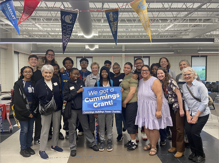Photo of alumni standing with alumni success coaches under a banner of college pennants holding a sign that says "We got a Cummings grant!"
